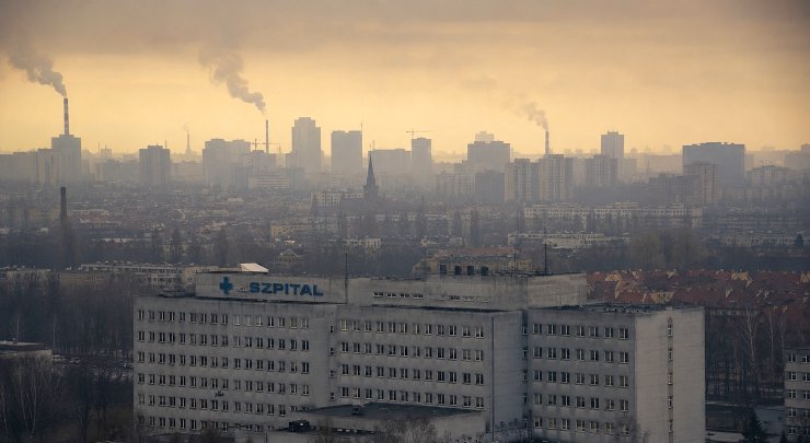 A photograph of a large concrete hospital with a blue SZPITAL sign against a hazy, smoggy city skyline with smoking chimneys